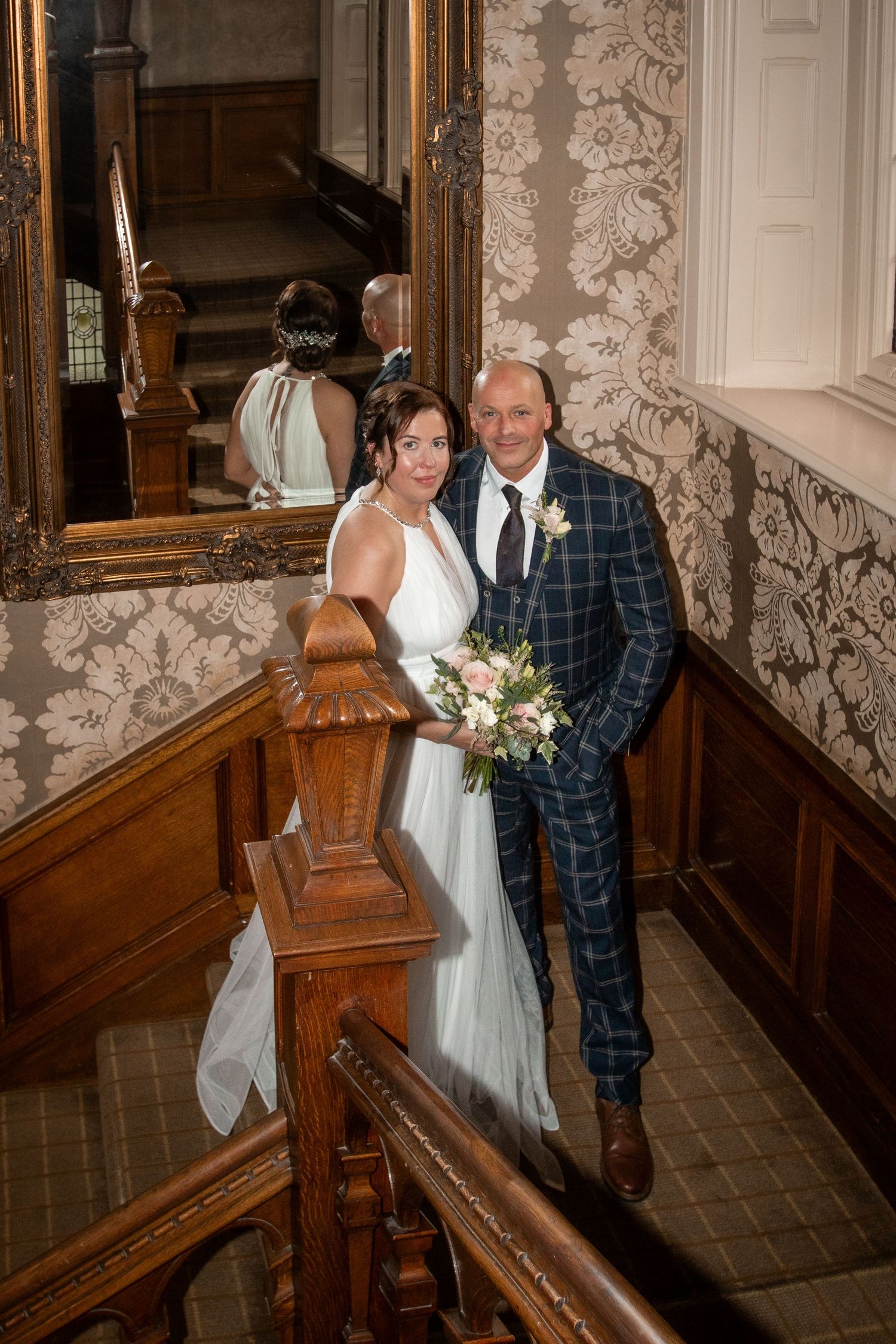 Bride and groom posing on wooden staircase in elegant venue, bride holding bouquet, groom in plaid suit, reflected in ornate mirror.