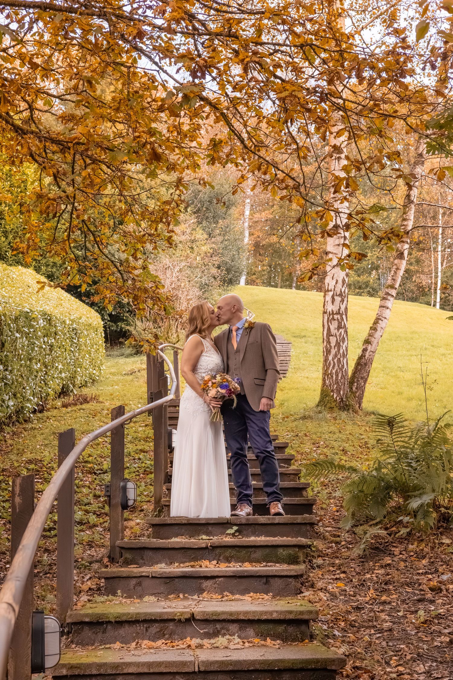 Newlyweds sharing a kiss on leaf-covered stairs beneath vibrant autumn trees in a scenic park.