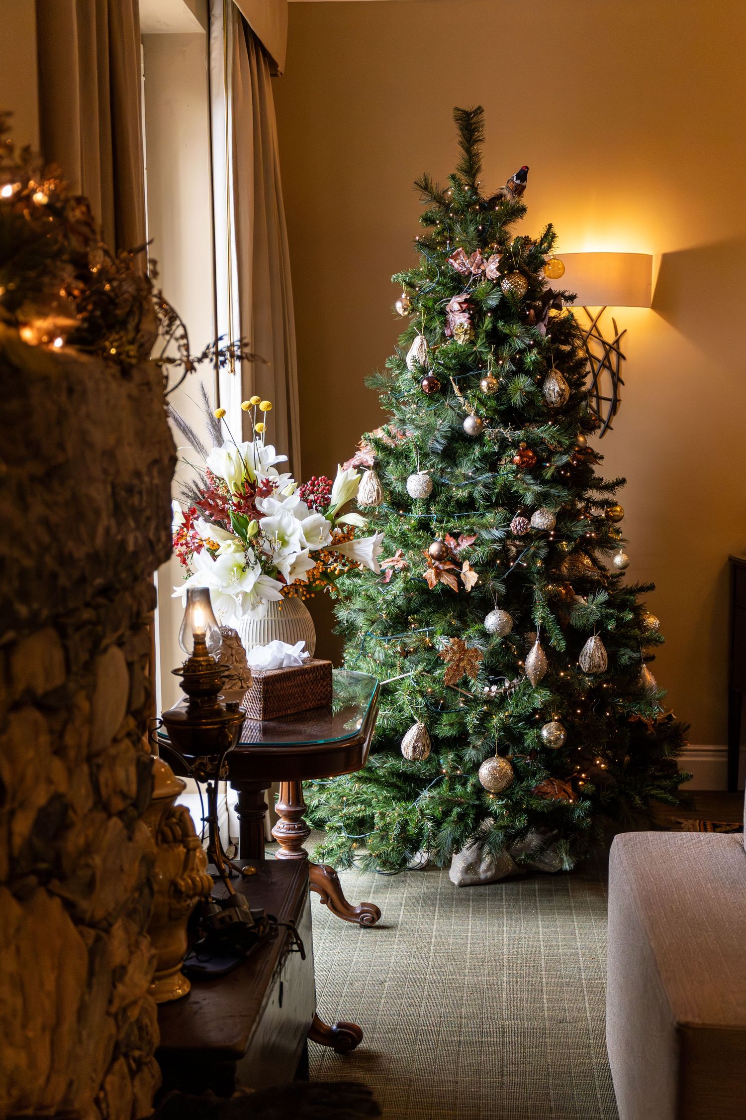 Elegantly decorated Christmas tree with ornaments beside a floral arrangement in a warm, cozy living room.