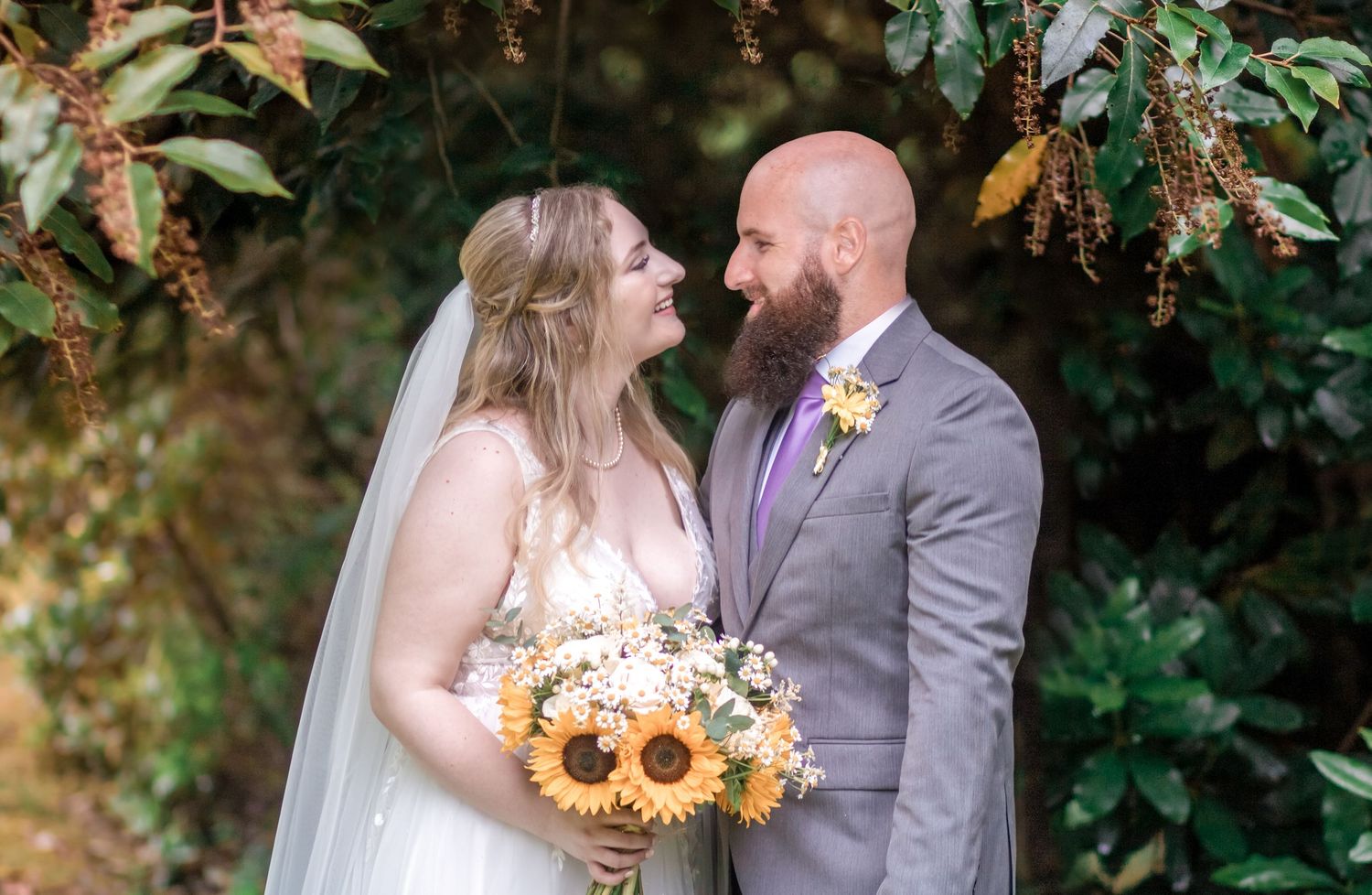 Bride and groom smiling at each other under floral arch, bride holding sunflower bouquet, groom in gray suit with purple tie.