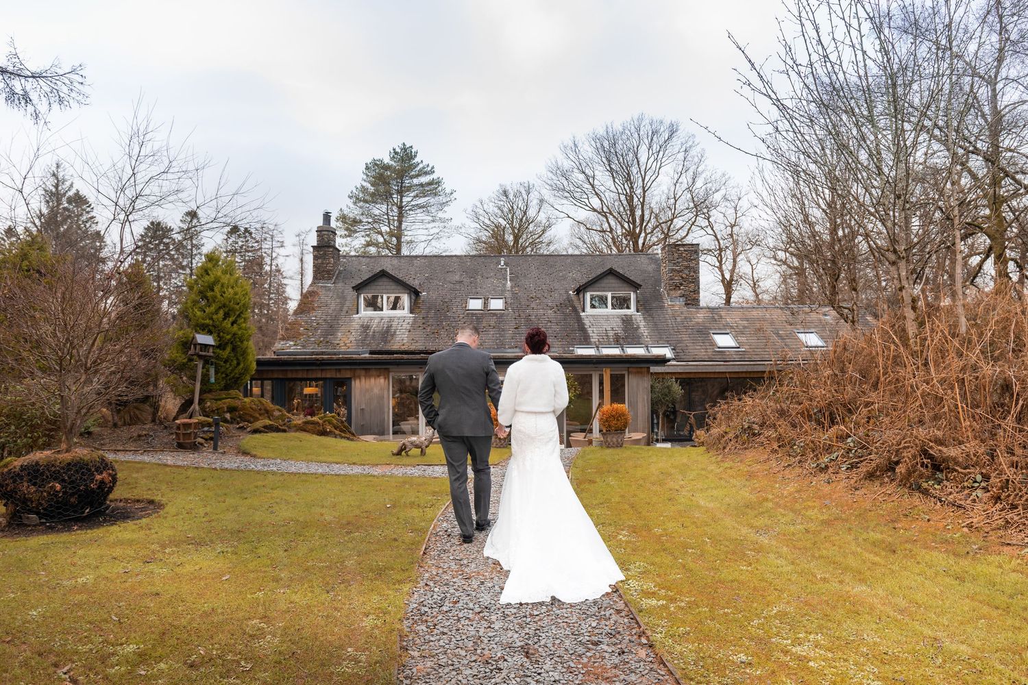 A bride and groom walk hand in hand toward an elegant country house surrounded by lush greenery on their wedding day.
