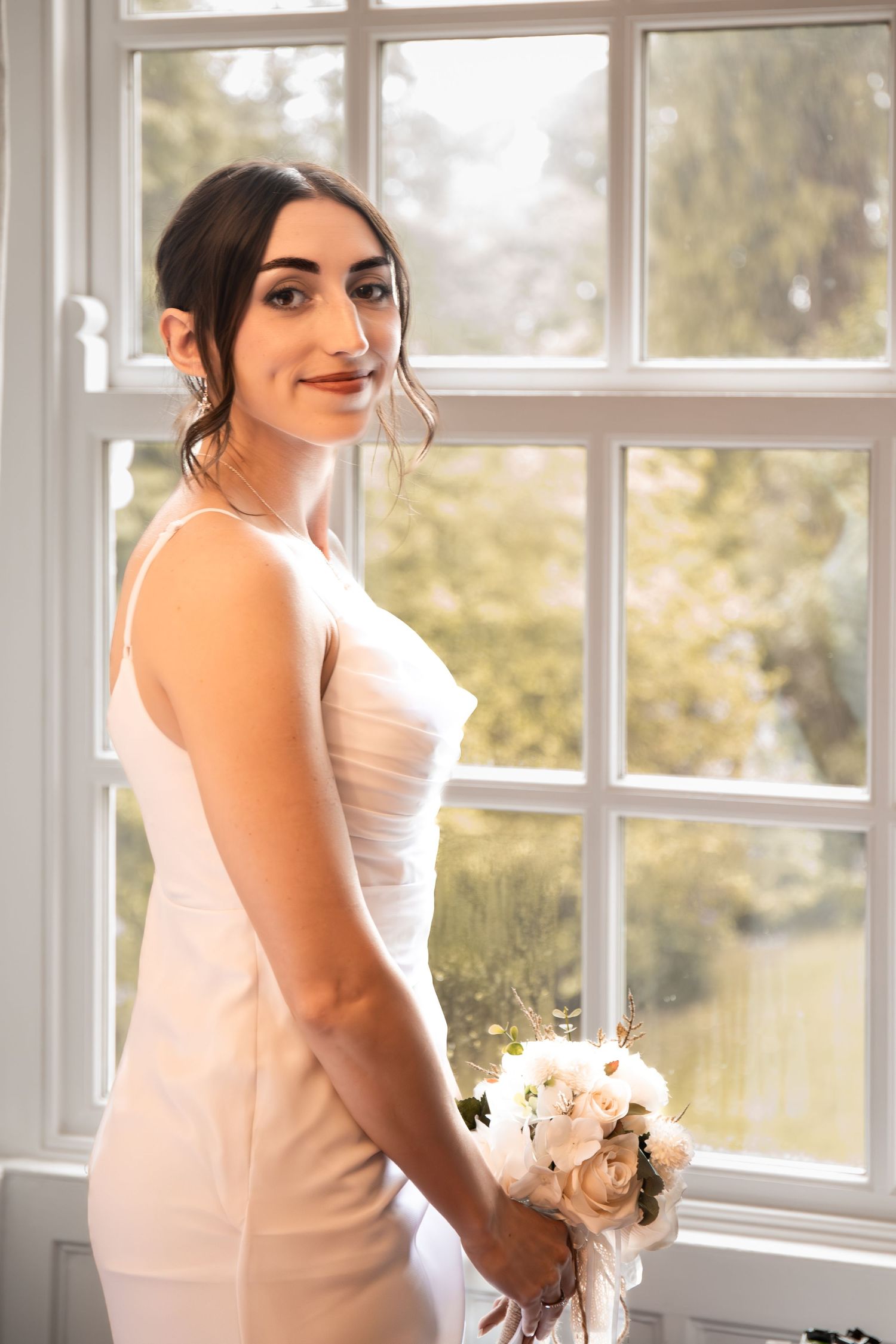 Bride in white dress holding bouquet of white flowers standing by large window with natural light.