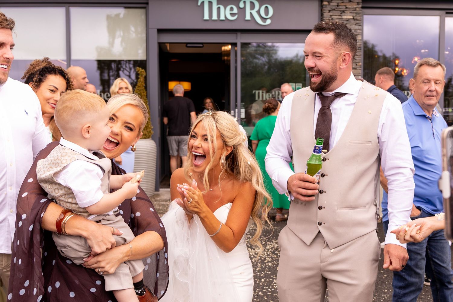 Bride and groom laughing with guests outside The Ro venue, bride in white dress, groom in grey suit holding beer.