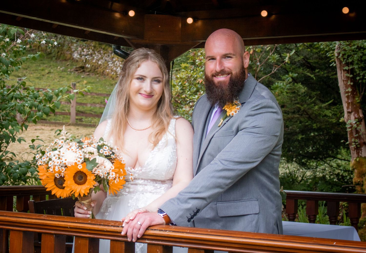 Bride holding sunflower bouquet and bearded groom in grey suit posing on a wooden deck in nature.