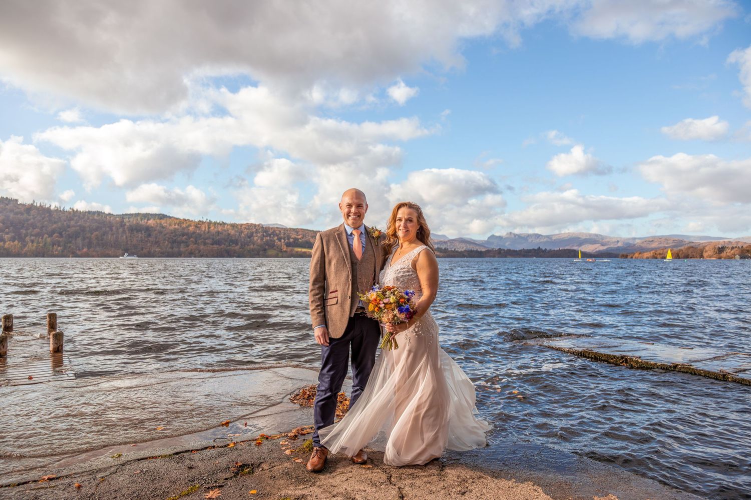 Bride in flowing gown and groom in tweed suit posing on rocky lakeside with autumn hills and cloudy sky behind.