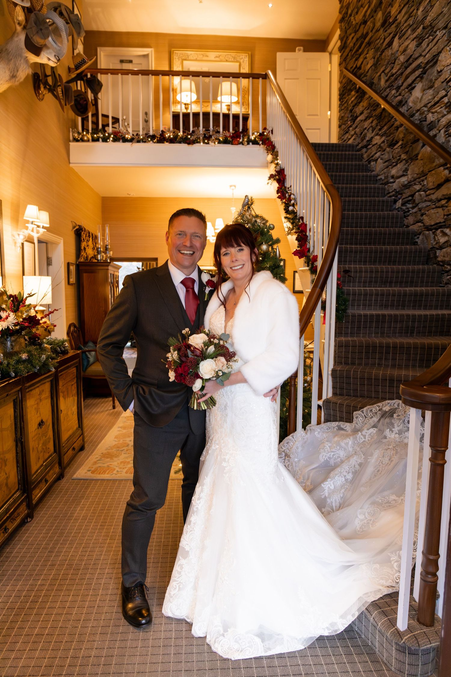 Bride and groom pose together in a decorated wedding venue hallway with a staircase and holiday greenery.