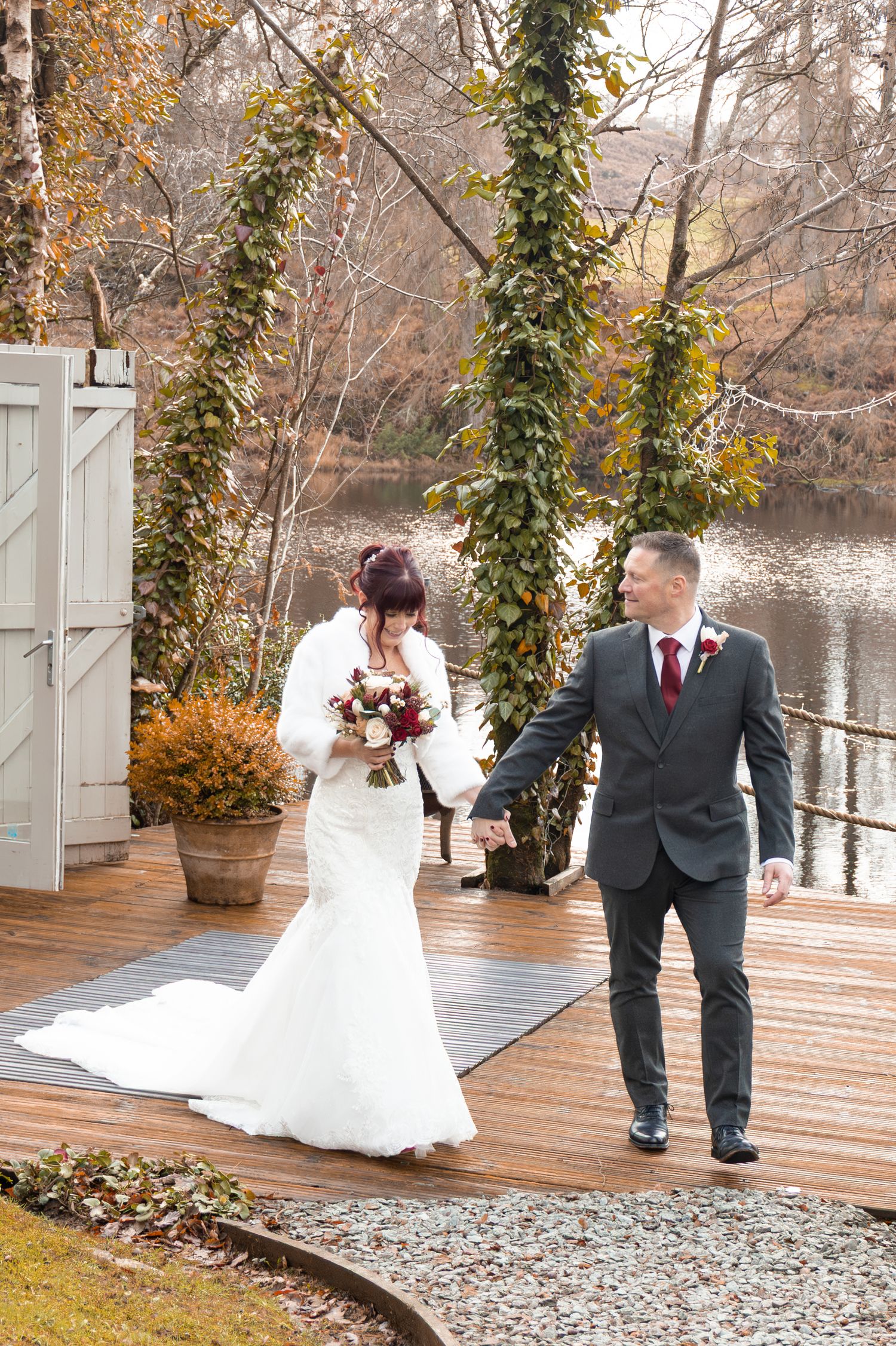 Bride and groom hold hands on a wooden deck beside a lake, surrounded by autumn foliage at their outdoor wedding.