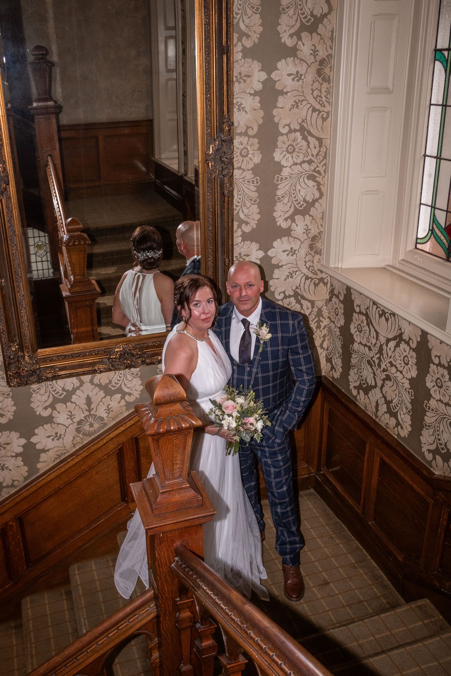 Bride and groom pose on ornate wooden staircase in elegant venue with gold mirror and stained glass window.