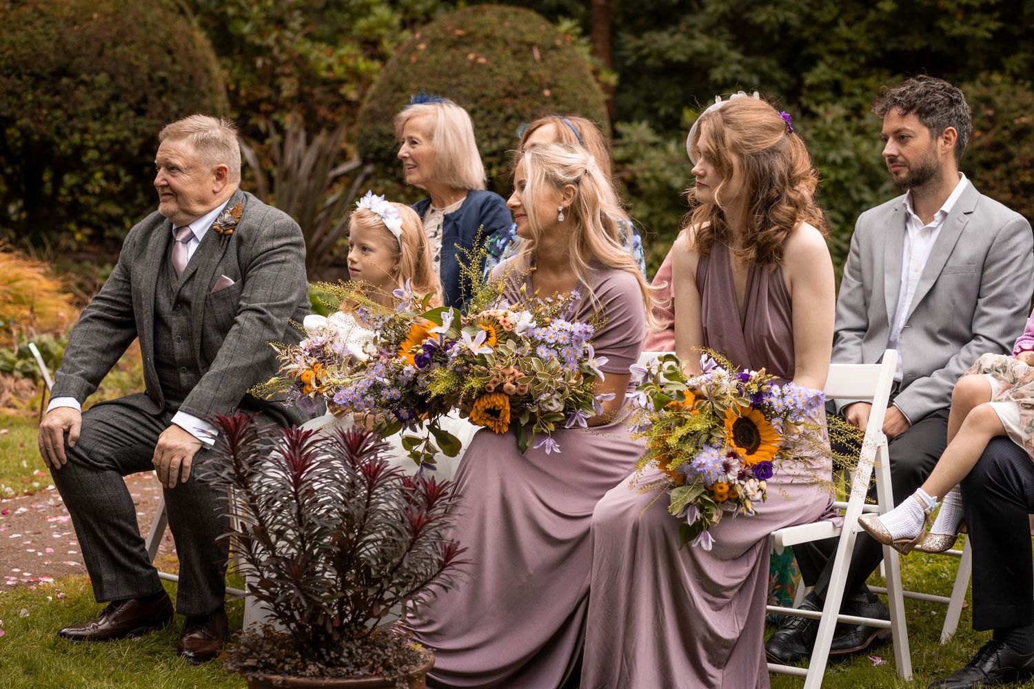 Bridesmaids in mauve dresses holding sunflower bouquets seated outdoors at a garden wedding ceremony.