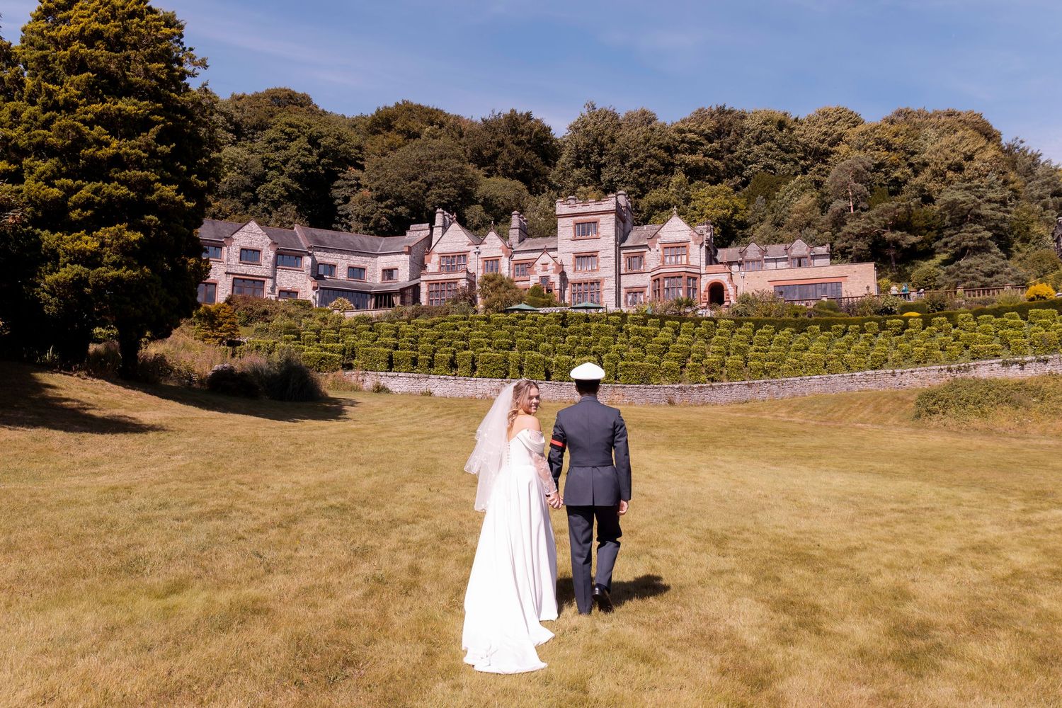Bride and groom walking hand in hand toward a grand manor house on a sunny wedding day.