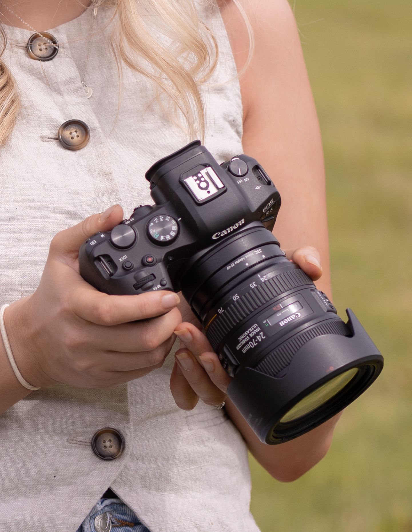 A woman holds a Canon DSLR camera with a zoom lens outdoors, wearing a linen button-up shirt.