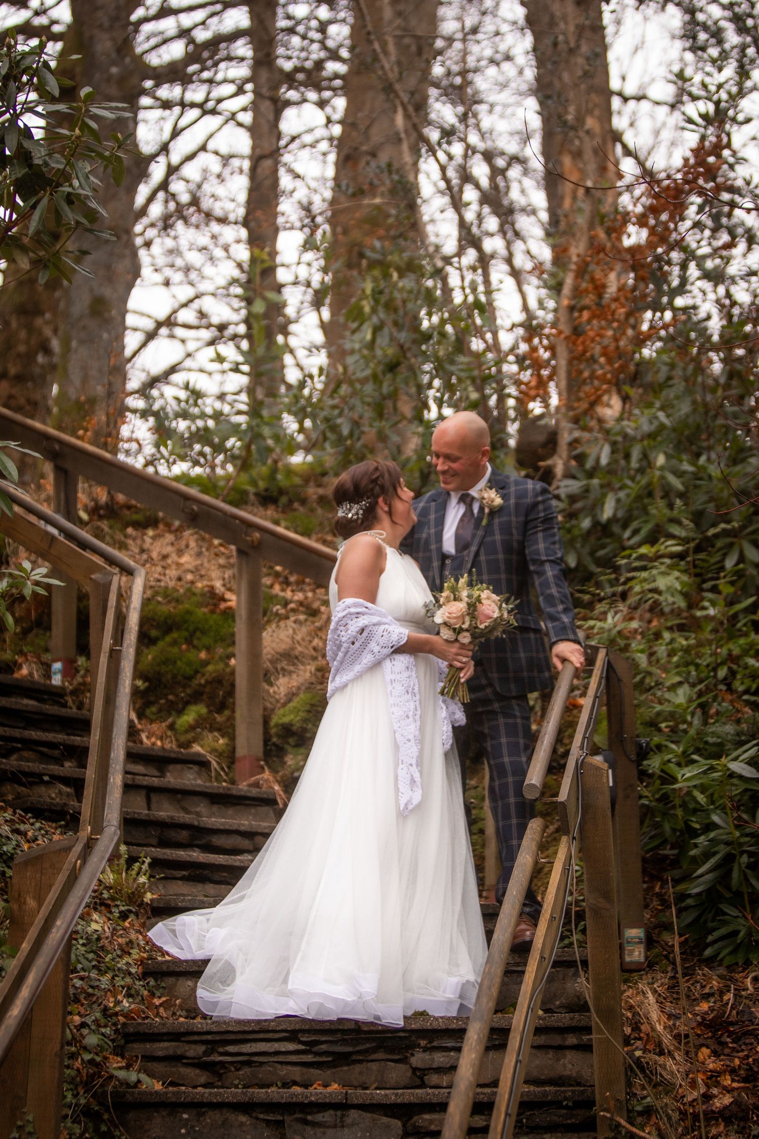 Bride and groom share a romantic moment on woodland steps, bride in white gown with bouquet, groom in plaid suit.