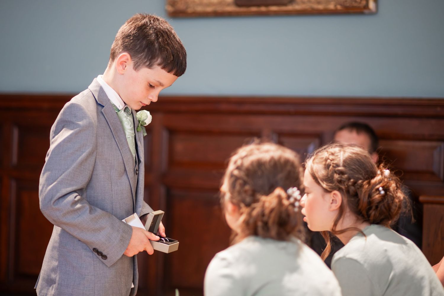 Young boy in gray suit holding wedding rings at a ceremony, with bridesmaids in mint dresses nearby.