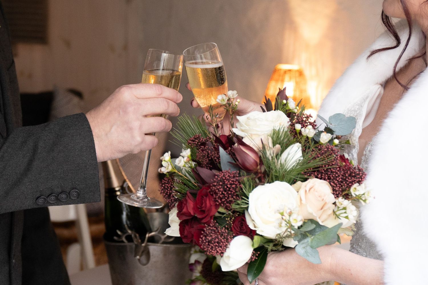 Bride and groom clink champagne glasses near a floral bouquet of red and white flowers at their wedding.