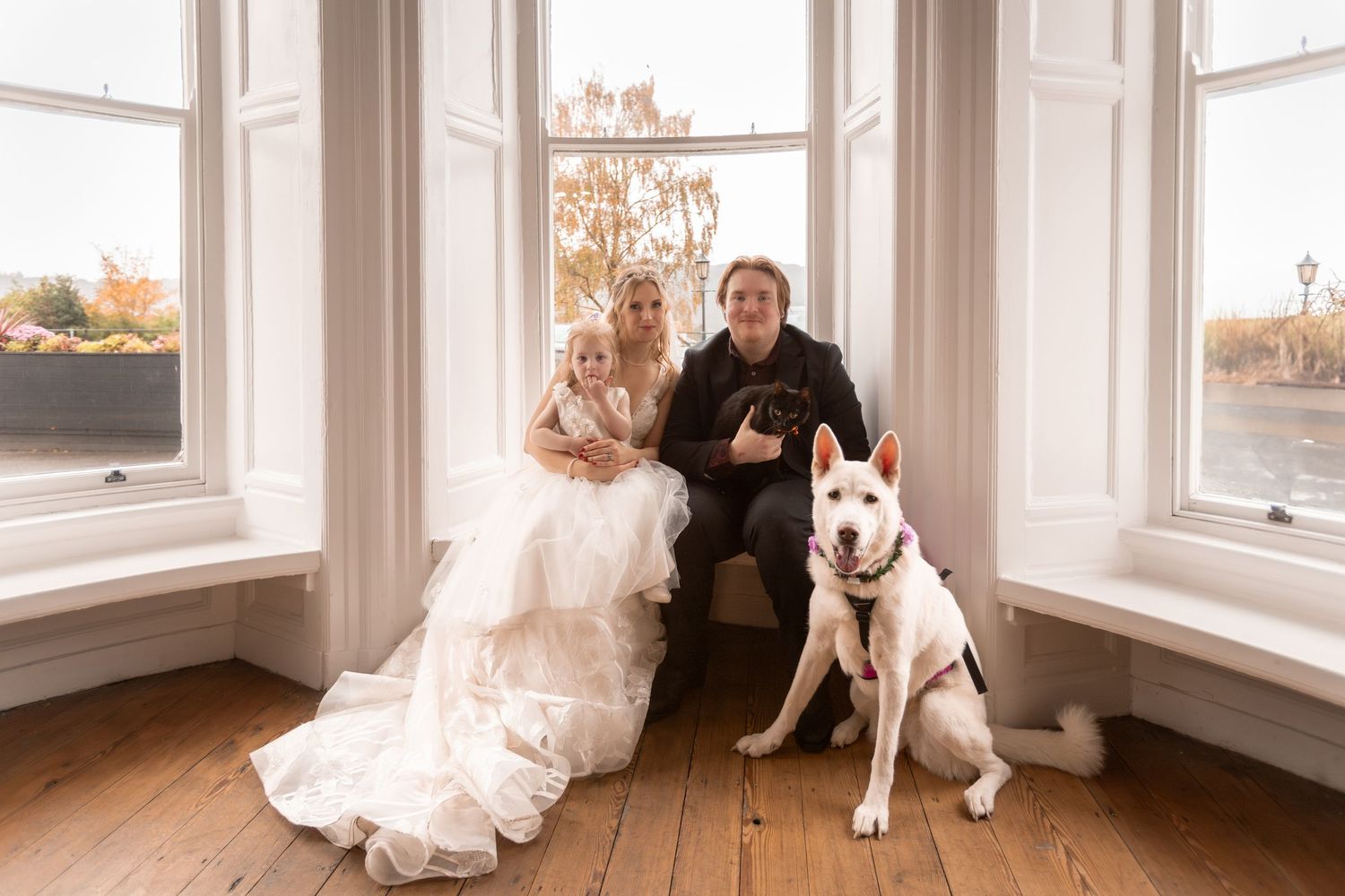 Bride and groom sit by bay windows with their white husky dog in a bright, elegant room.