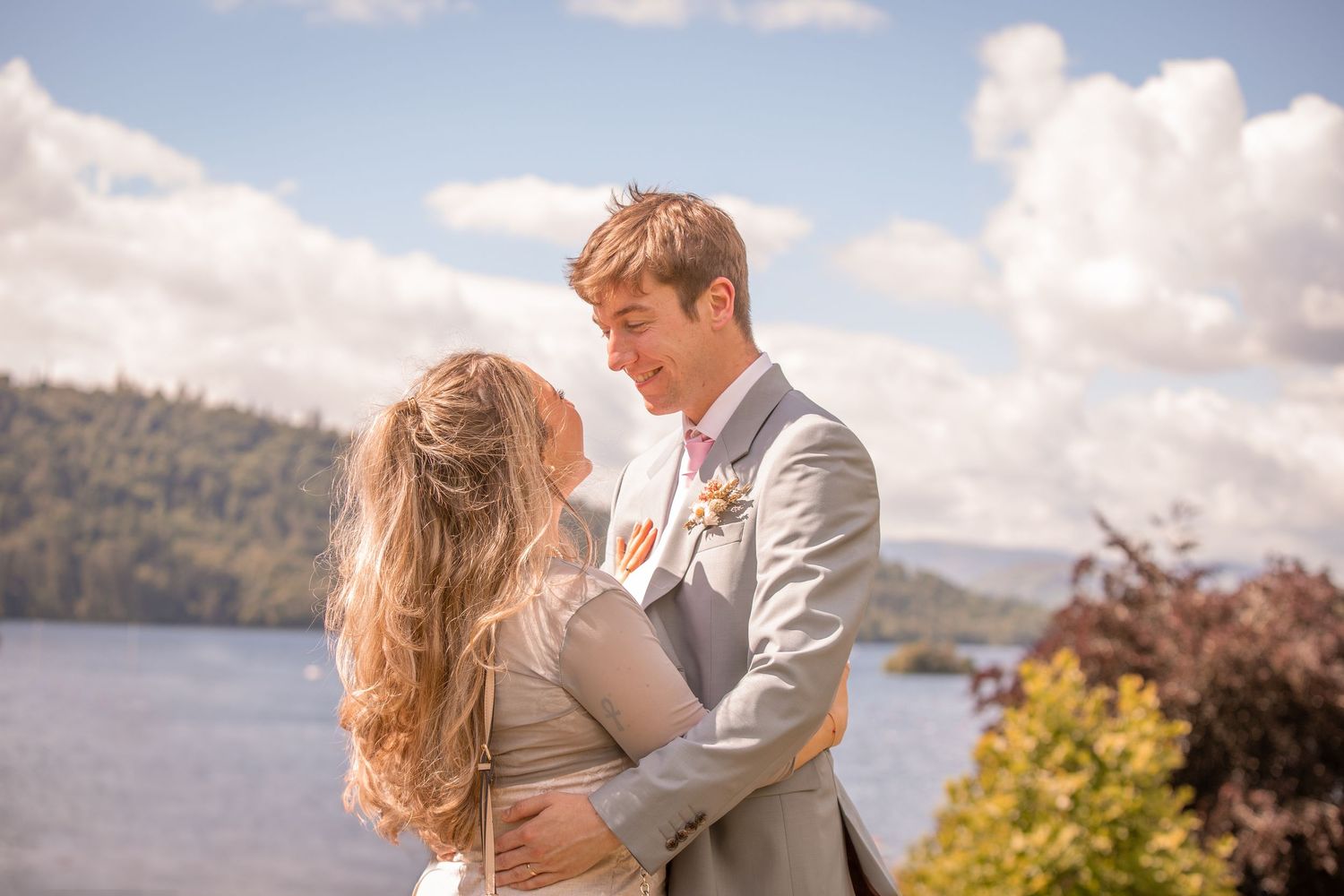 Romantic couple embracing outdoors near a lake, groom in grey suit, bride with flowing hair under blue sky.