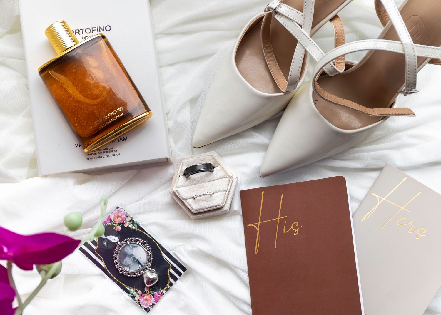Elegant wedding flat lay with perfume bottle, white heels, ring box, watch, and His journal on white fabric.