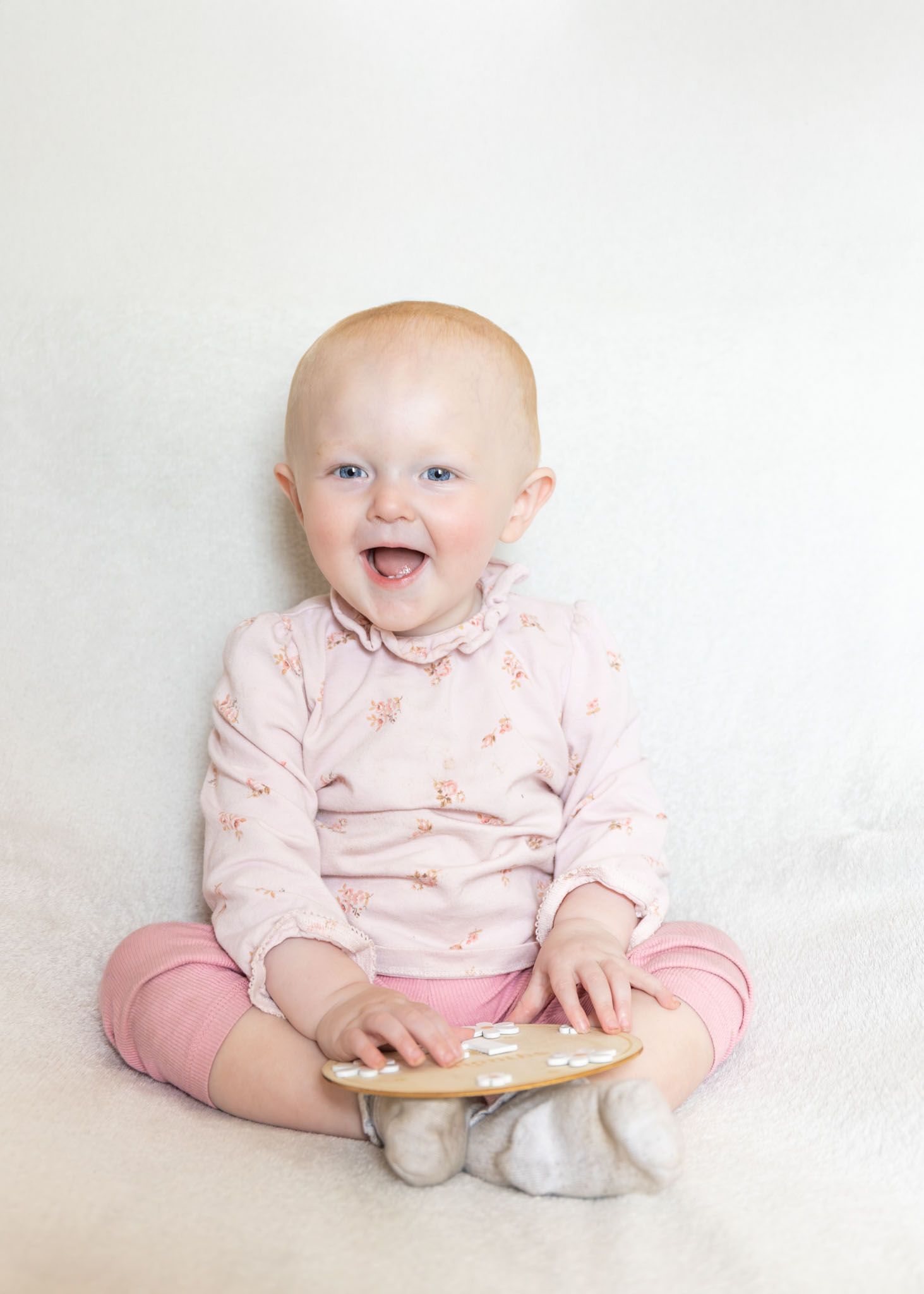 Smiling baby girl in pink floral outfit sitting upright on white fluffy blanket, holding a small wooden toy.