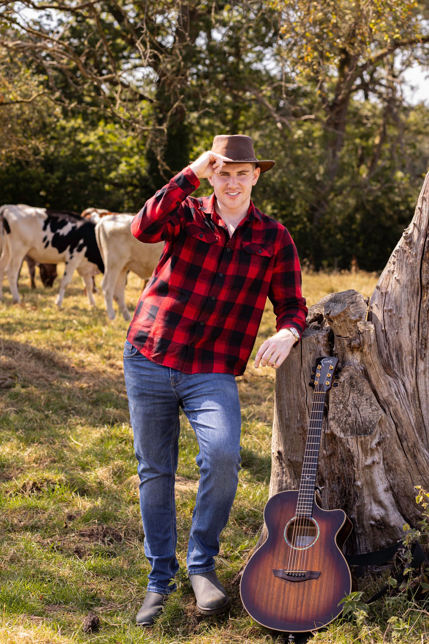 Young man in red plaid shirt and cowboy hat leans on tree stump with acoustic guitar, cows grazing behind him.