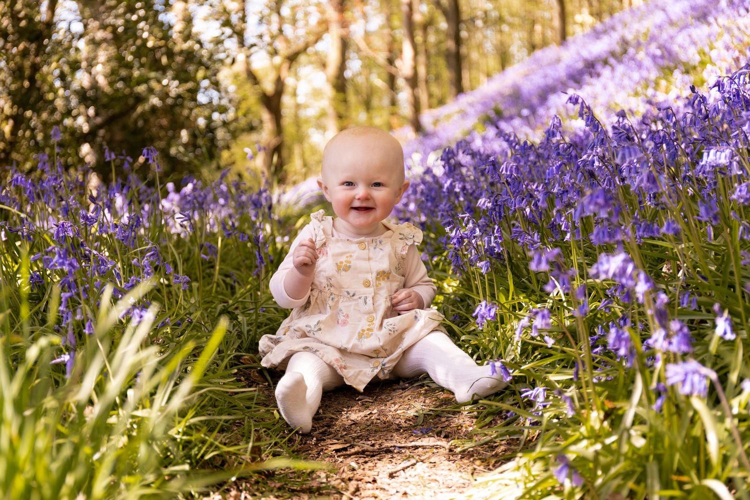 Happy baby sitting among vibrant purple bluebells in a sunlit woodland forest path.