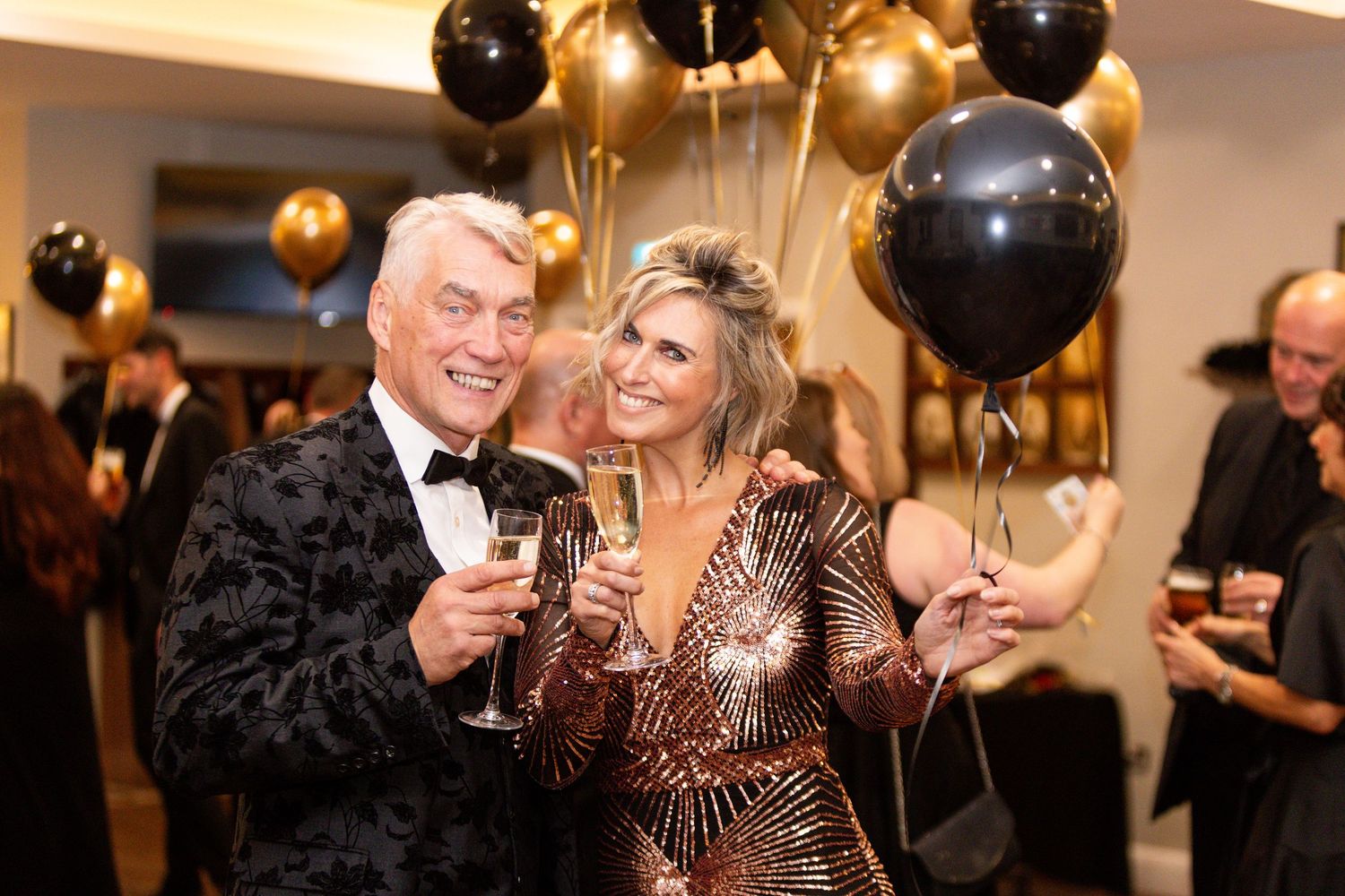 Elegant couple toasting with champagne glasses at a glamorous New Year's Eve party with gold and black balloons.