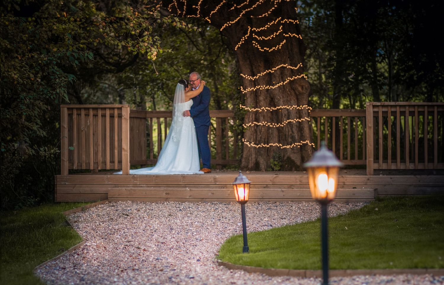 Romantic couple embracing on a wooden deck surrounded by fairy lights and lanterns at night.