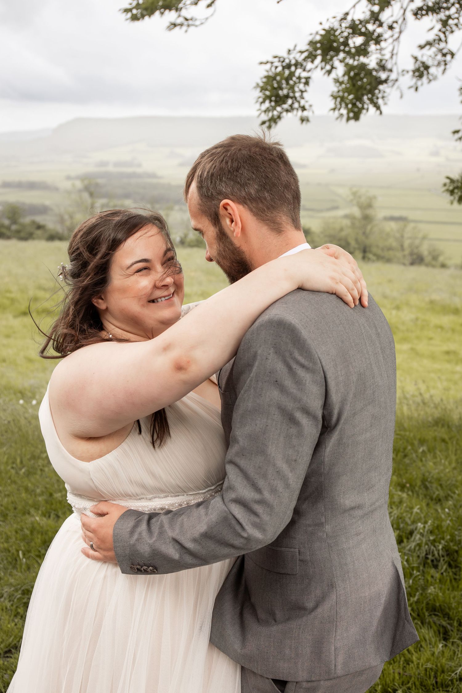 Smiling bride in white gown embraces bearded groom in gray suit outdoors on a green hillside.