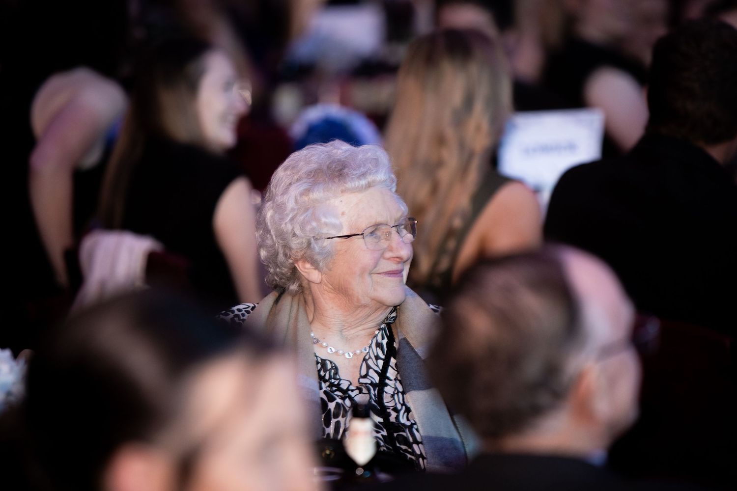 Elderly woman with white hair and glasses smiling warmly at a formal evening event.