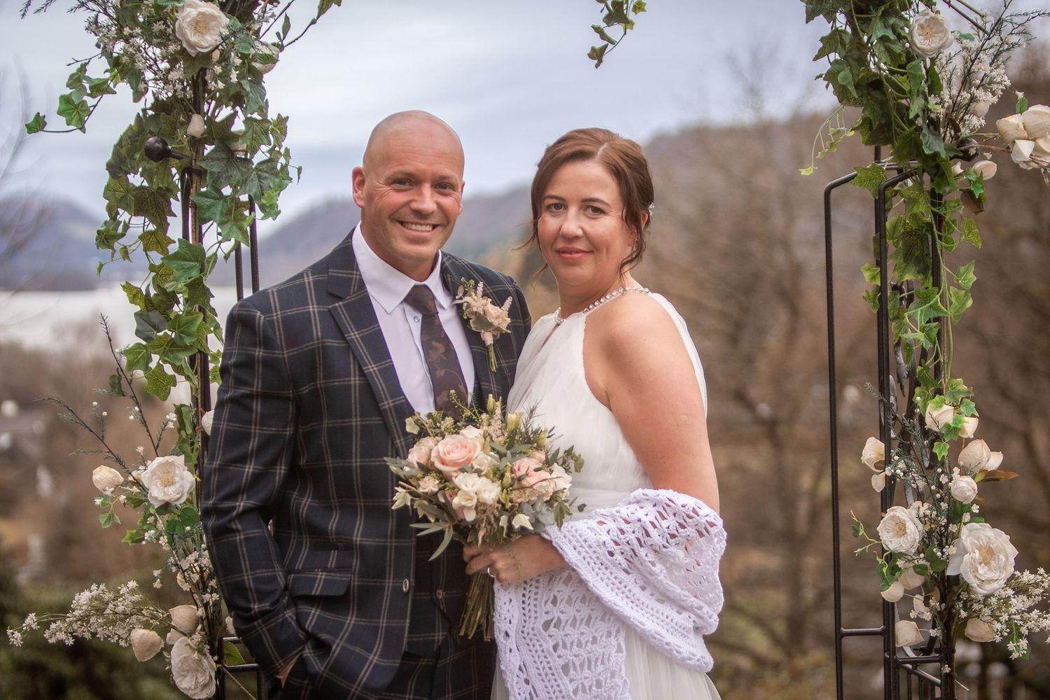 Smiling bride and groom posing together under a floral arch adorned with white roses and greenery.