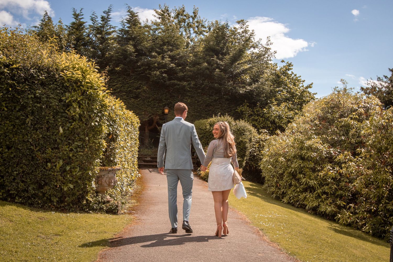 Bride and groom walking away hand in hand down a garden path lined with hedges on their wedding day.