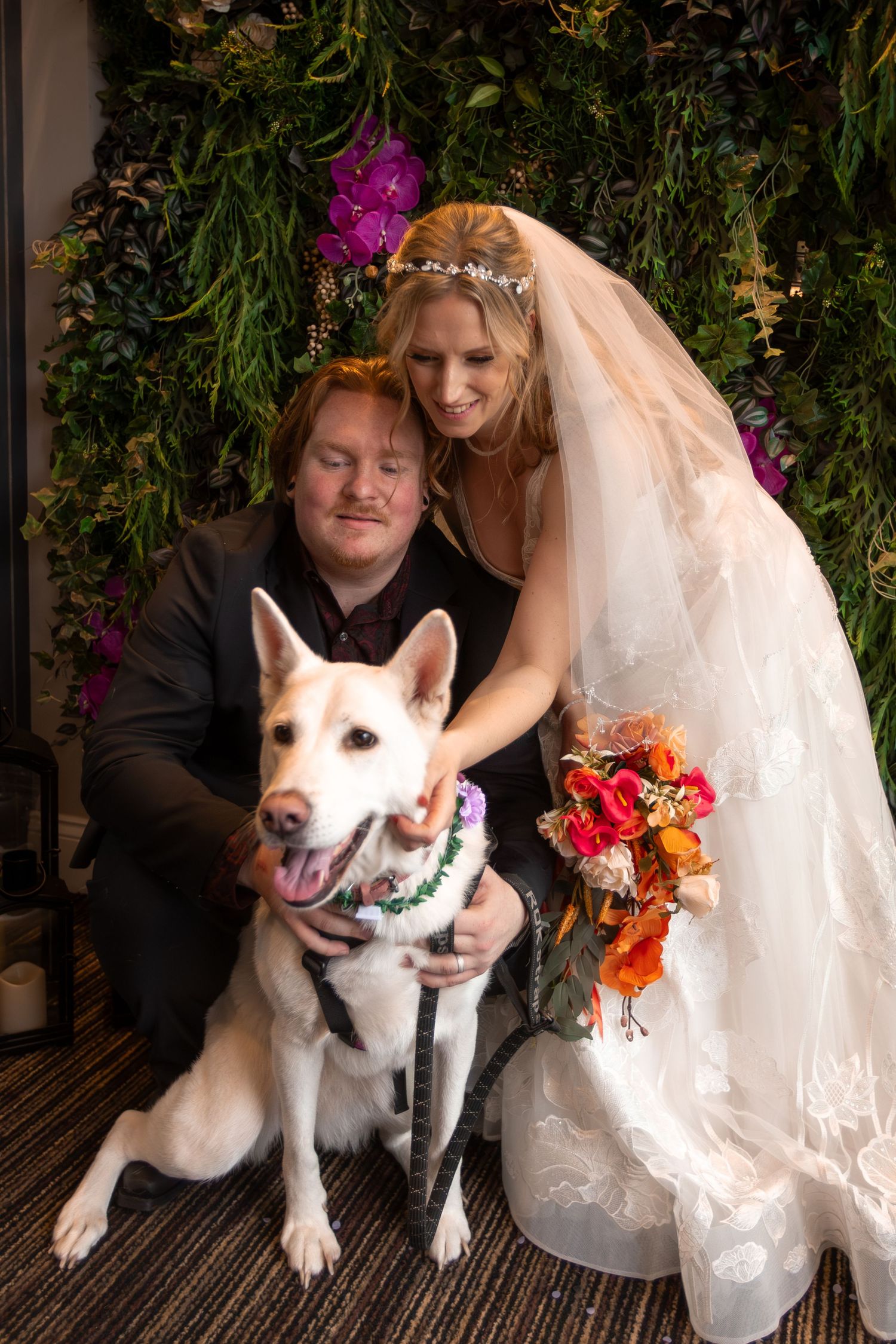 Bride and groom posing with their white dog wearing a floral collar in front of a lush green floral backdrop.