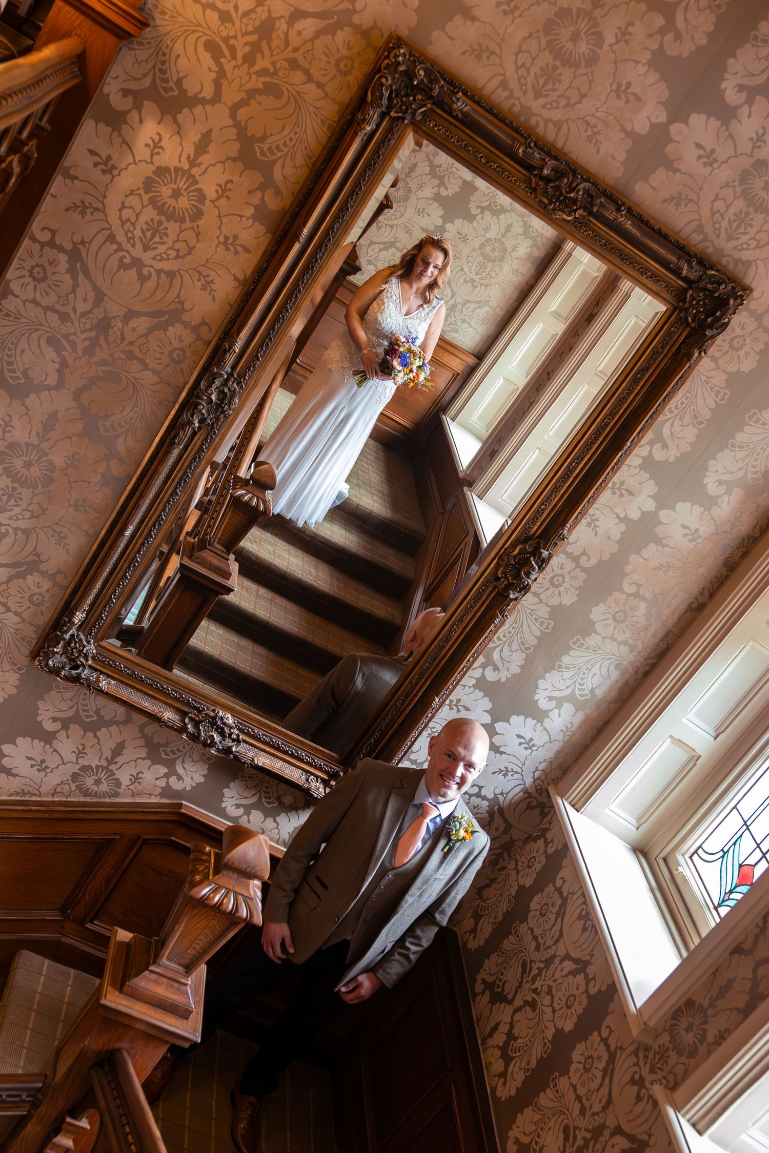 Creative wedding photo using ornate mirror reflection showing bride on staircase with groom below.