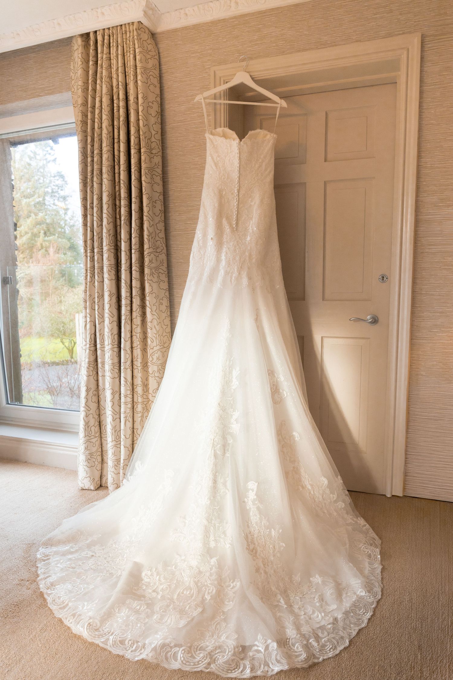 Elegant strapless lace wedding dress with long train hanging on door in bright bridal suite room.