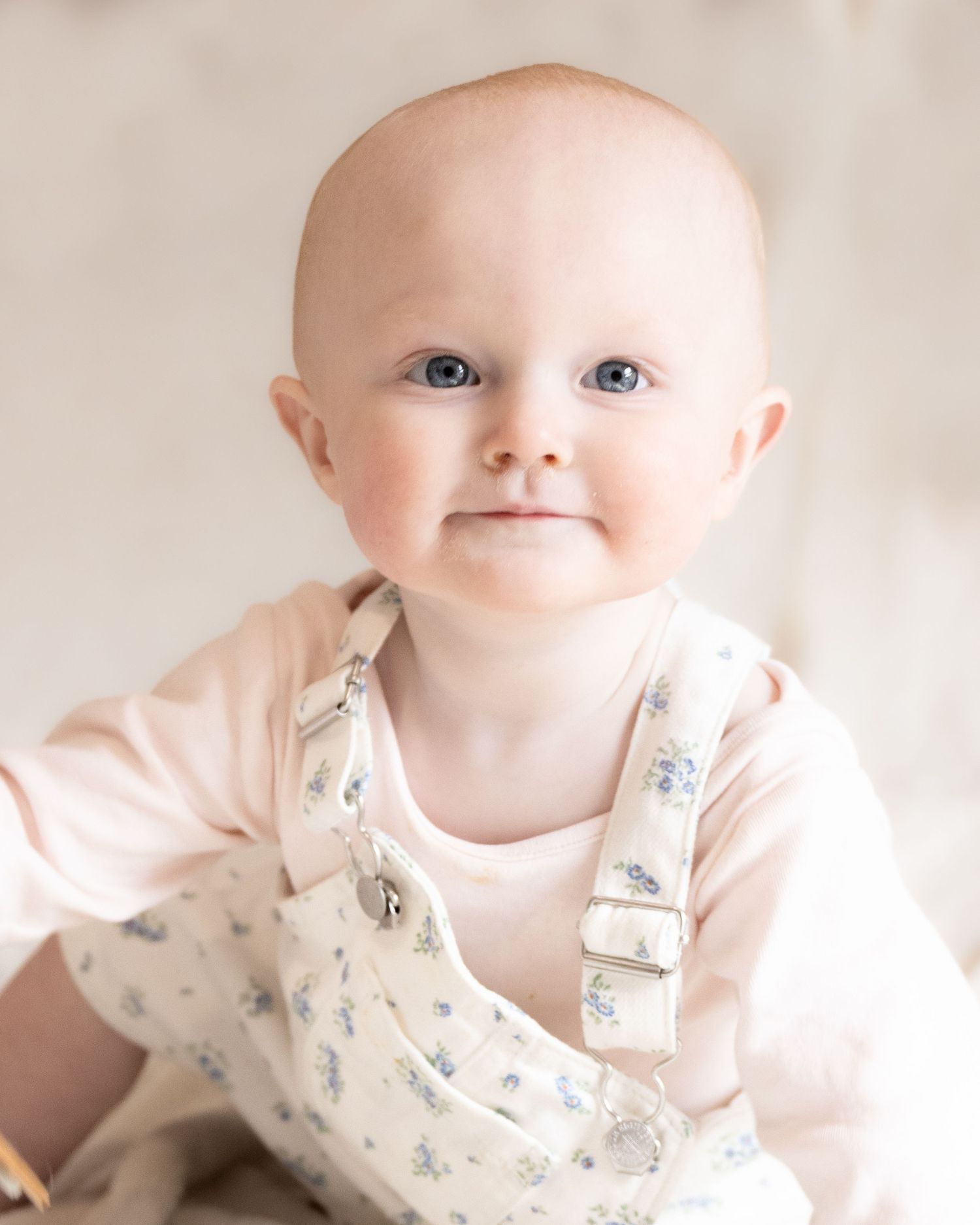 Adorable baby with blue eyes wearing floral overalls and pink top in a soft studio portrait.