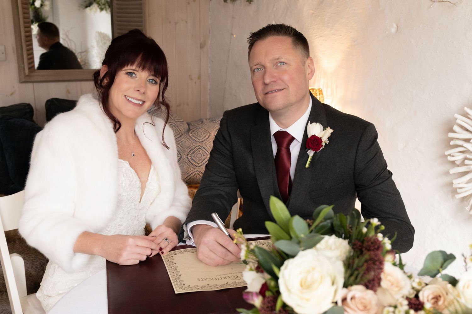 Smiling bride in white fur wrap and groom signing wedding register at decorated table.