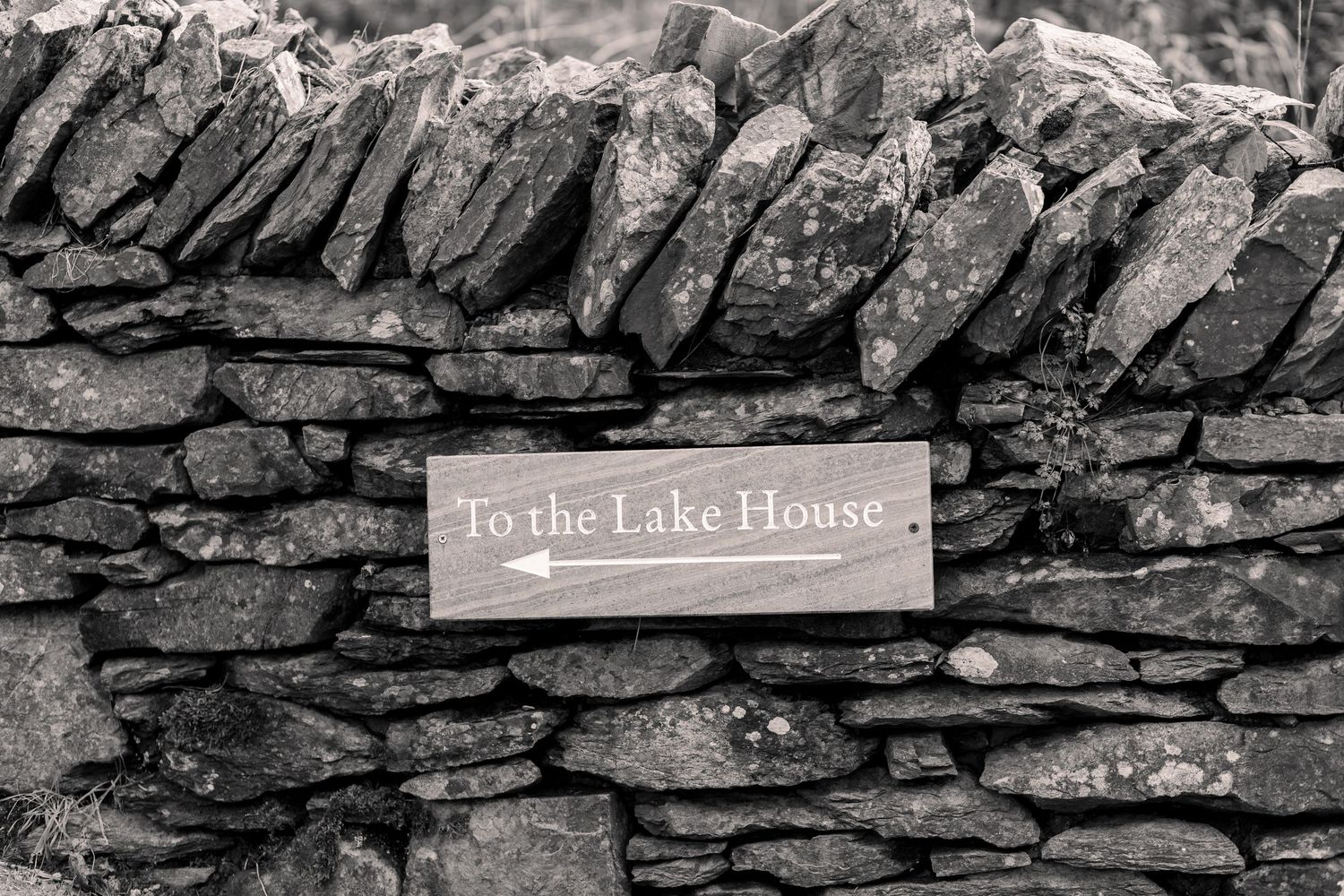 Black and white photo of a wooden directional sign reading To the Lake House mounted on a rustic dry stone wall.