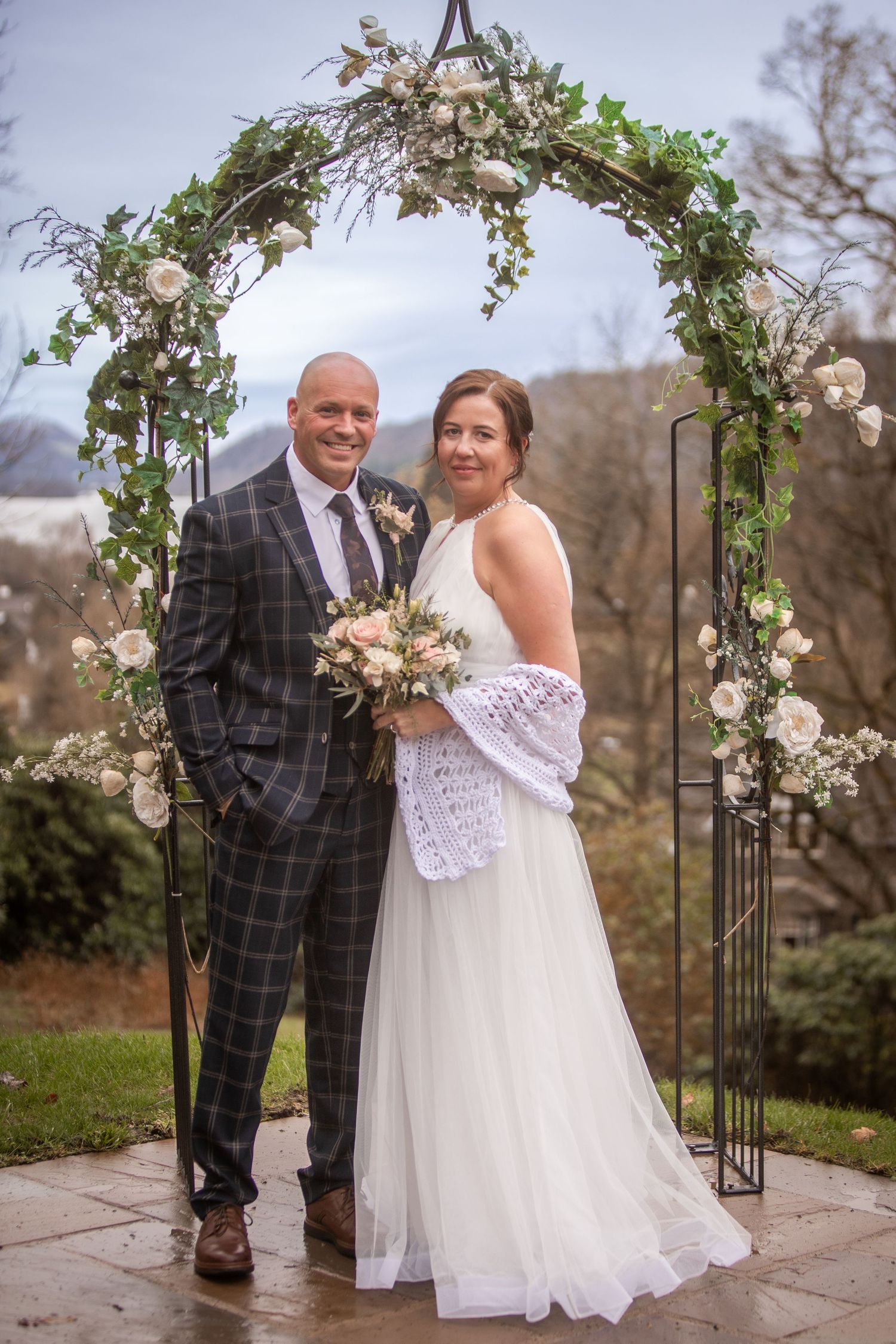 Bride and groom posing under a floral arch with greenery and white roses on their wedding day.