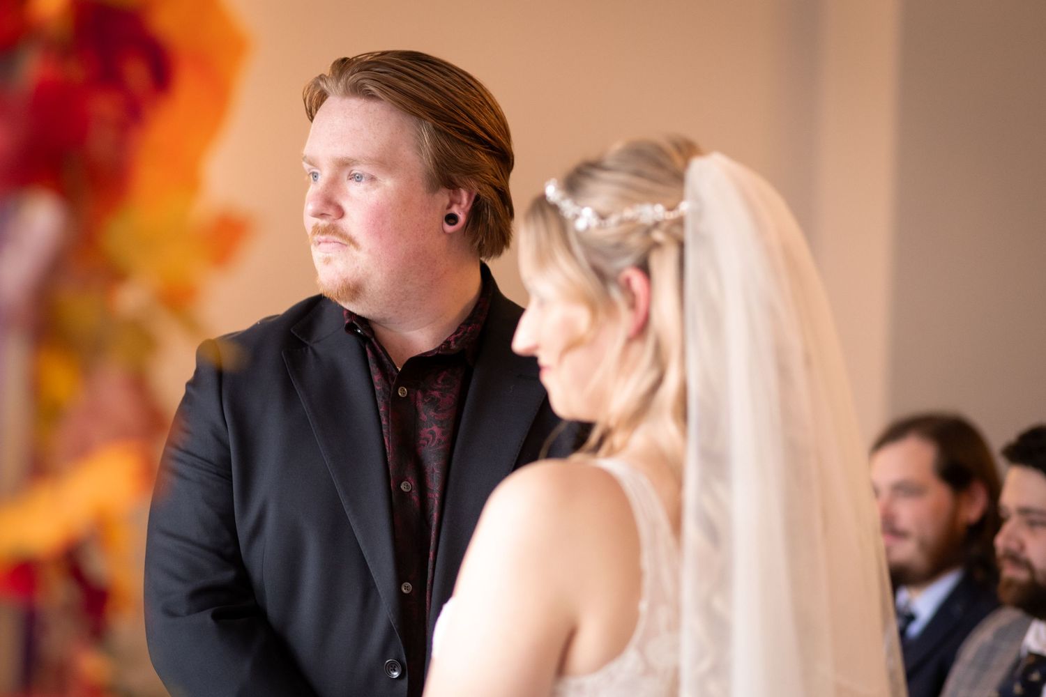 Groom in black suit watches bride in white veil during indoor wedding ceremony with autumn floral decor.