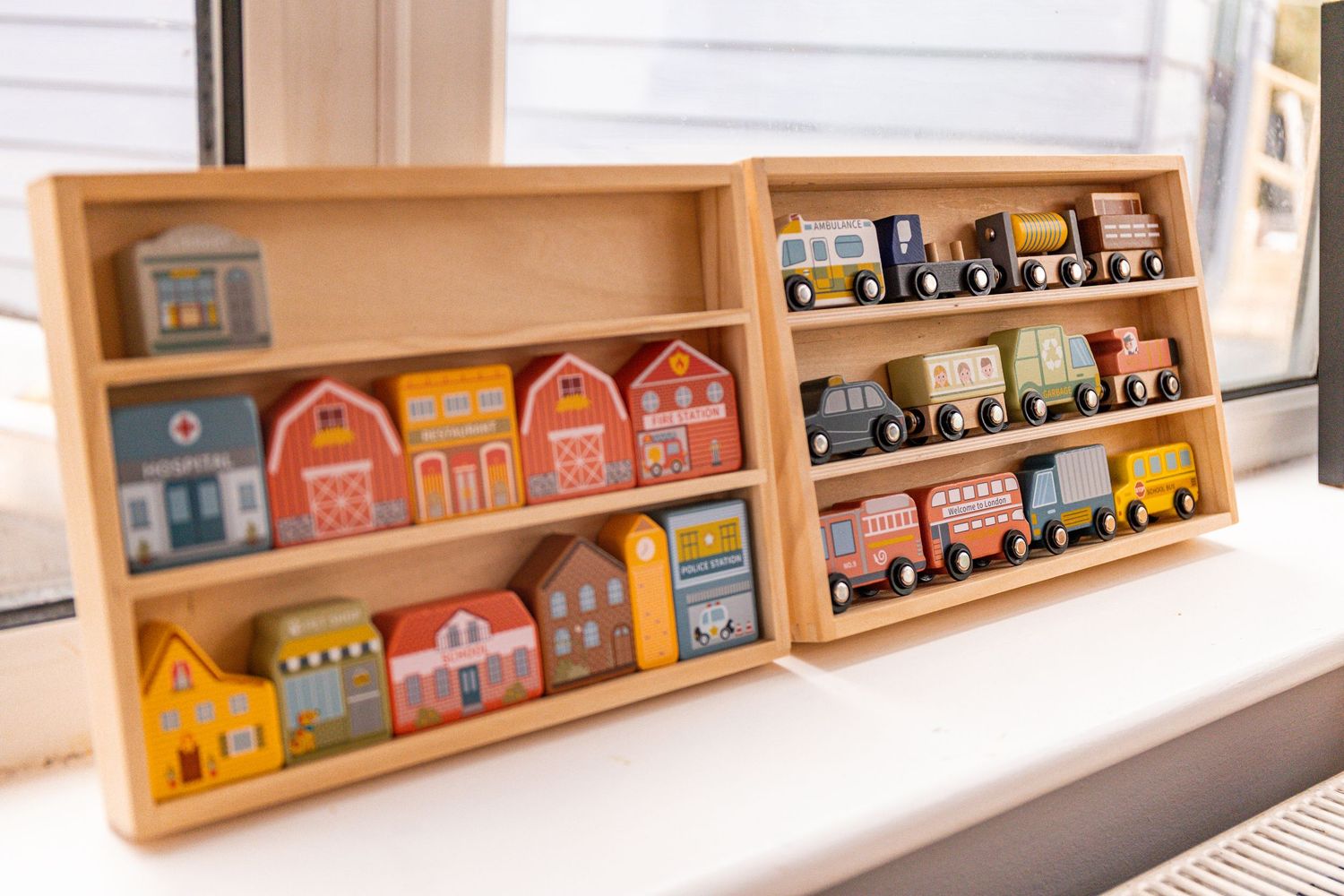 Colorful wooden toy village buildings and train set displayed on two wooden shelf units by a window.