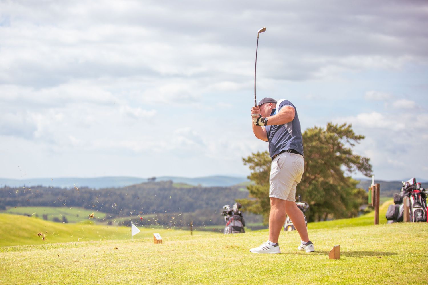 Male golfer finishing a drive shot on a sunny golf course with scenic hills.