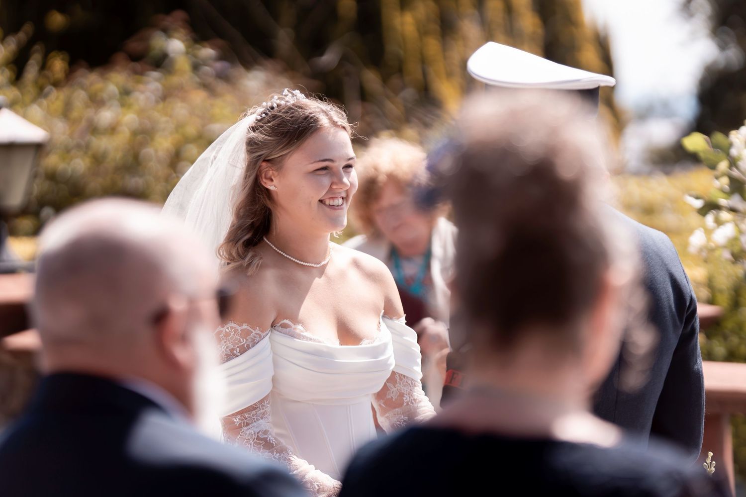 Smiling bride in white off-shoulder gown and veil during outdoor wedding ceremony on a sunny day.