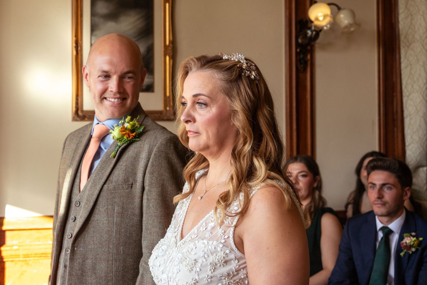 Bride and groom standing together during wedding ceremony, bride in lace dress with floral hairpiece, groom in grey suit.