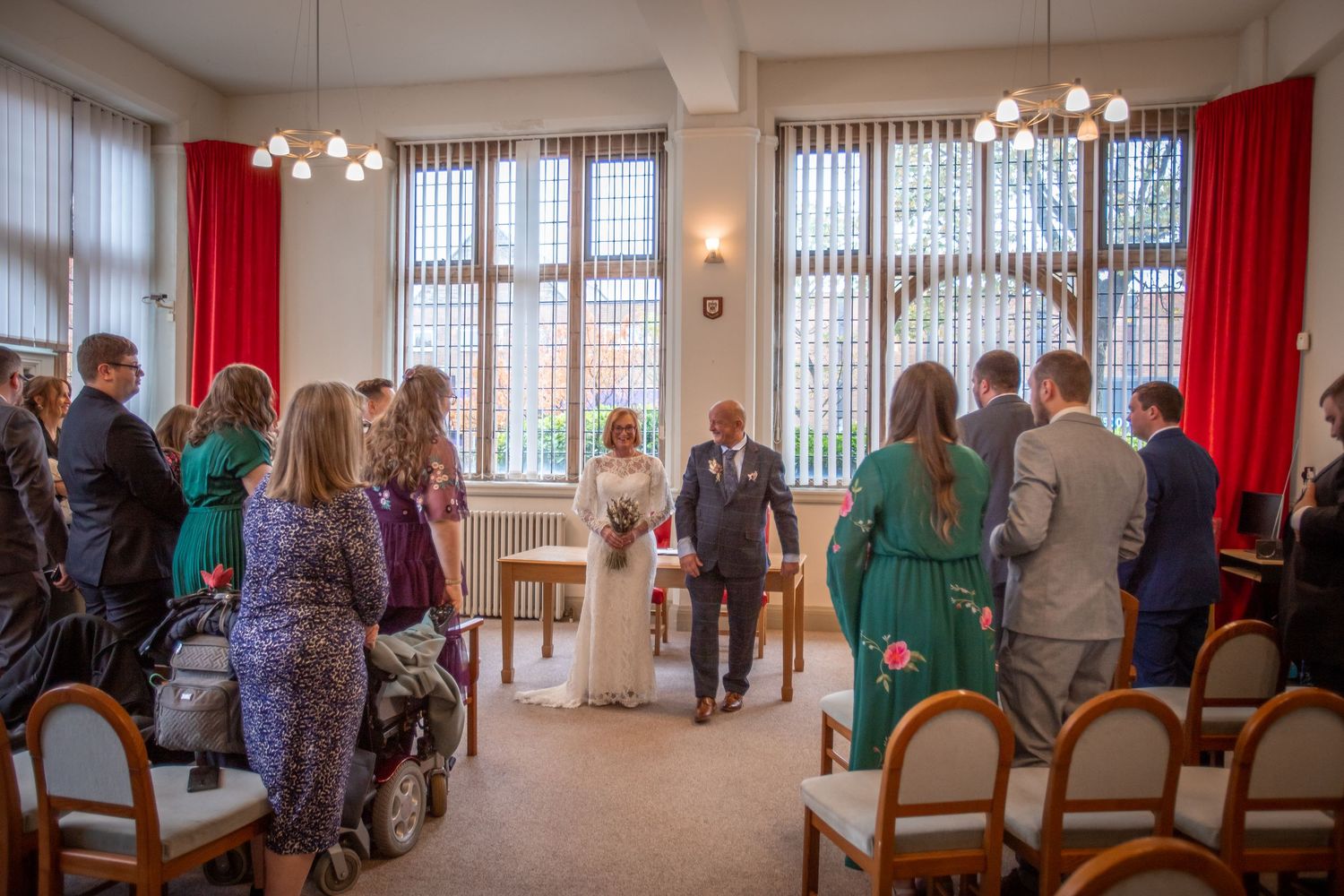 Guests gather for an intimate wedding ceremony in a bright room with red curtains as the bride and groom stand at the altar.