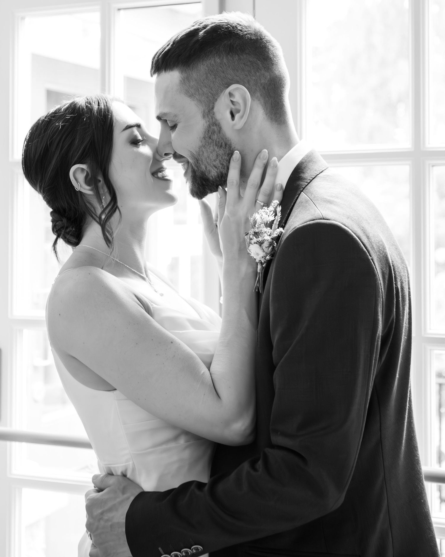 Black and white wedding photo of bride and groom sharing a romantic moment, nearly kissing by a bright window.