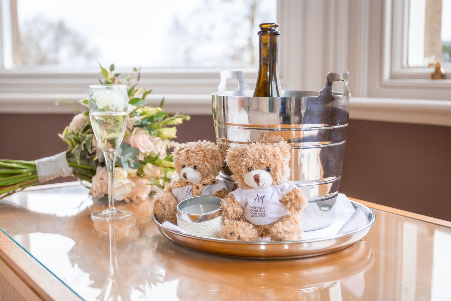 Two teddy bears on a silver tray with champagne flutes, a bottle in an ice bucket, and flowers on a wooden table.