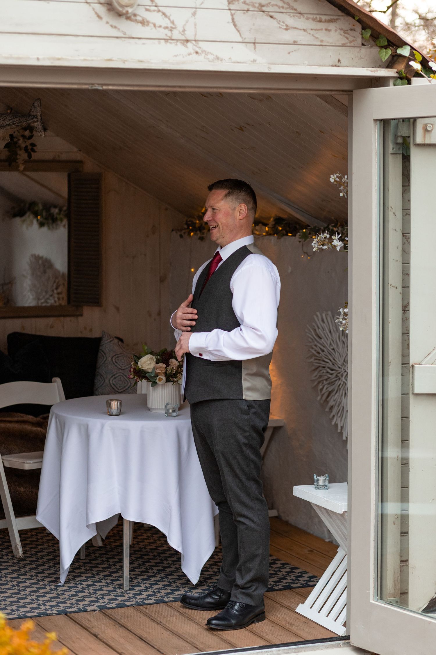 Groom in grey vest and red tie smiling in a decorated wedding venue doorway.
