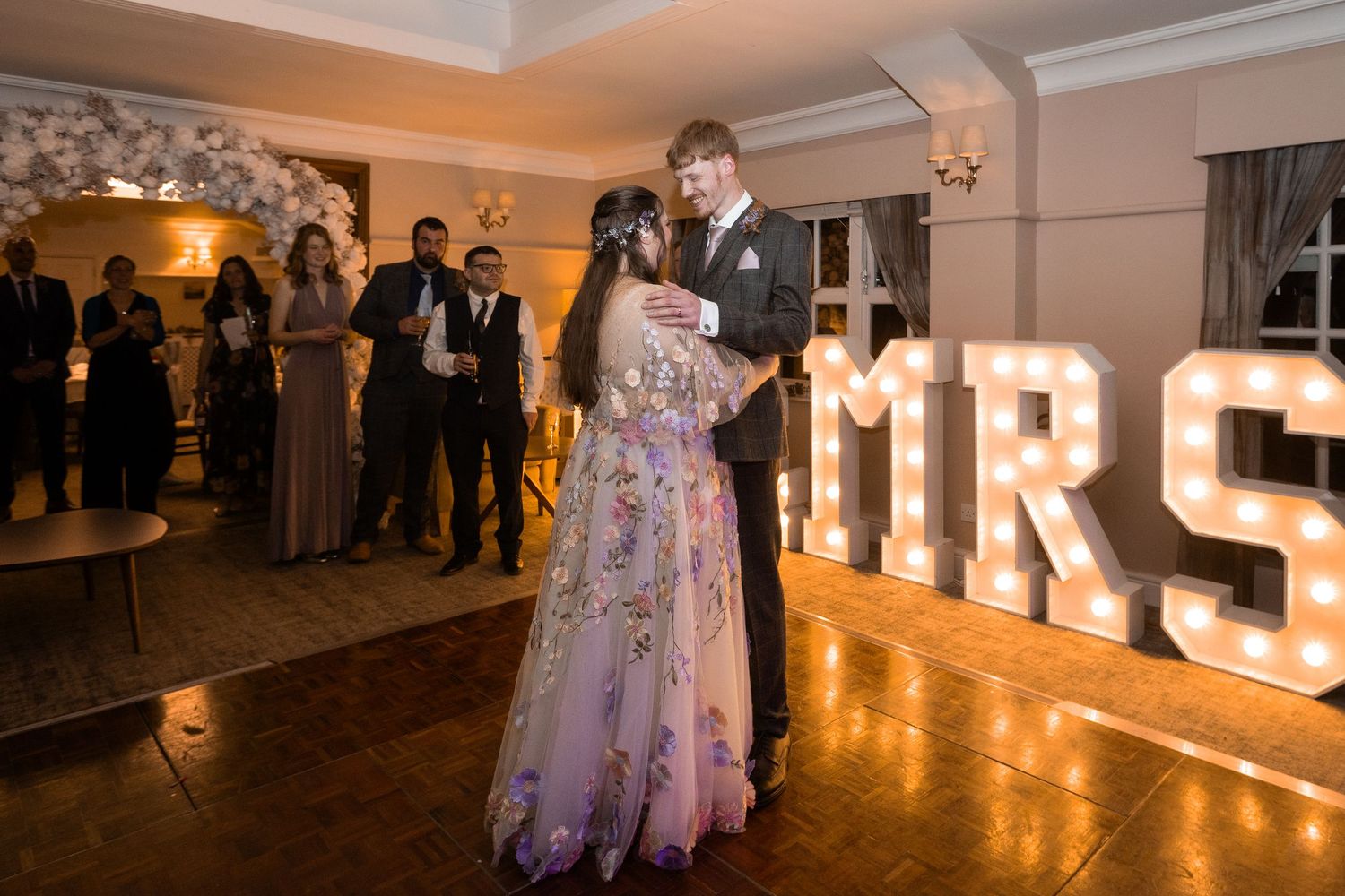 Couple sharing first dance at wedding reception with illuminated MRS marquee letters and guests watching.