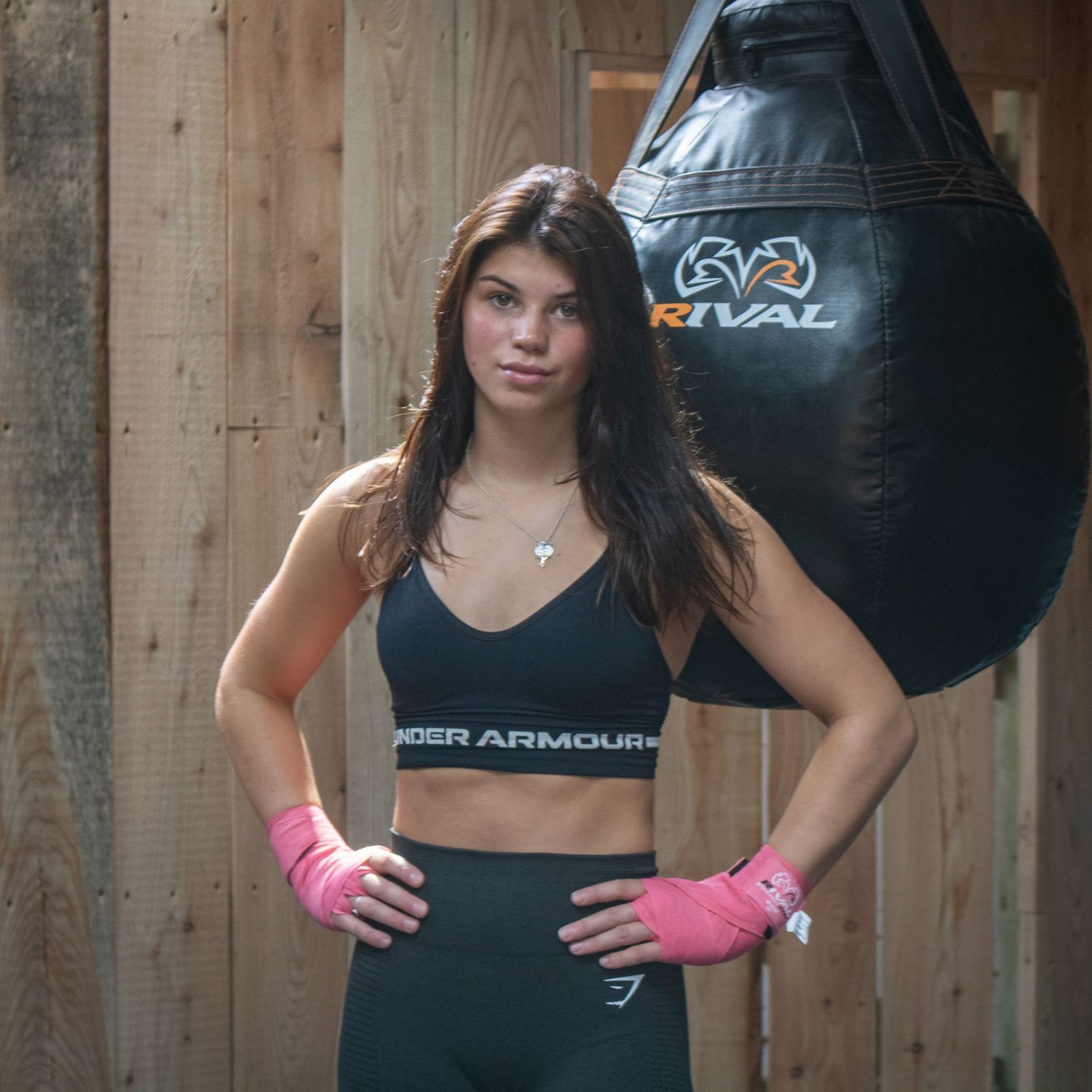 Female boxer in Under Armour sports bra with pink hand wraps posing beside a Rival heavy bag.