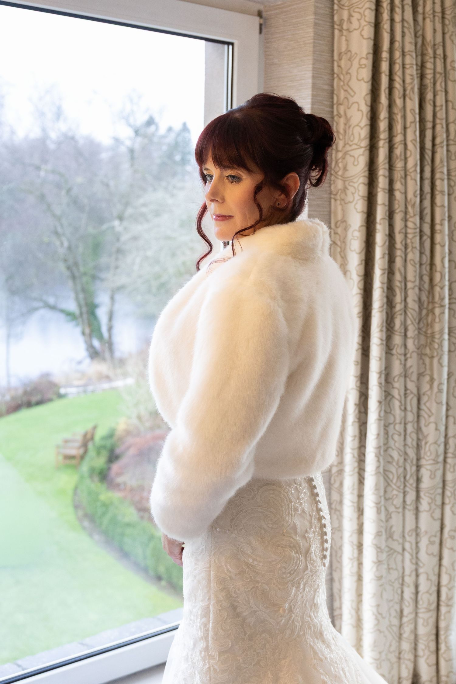 Elegant bride in white lace gown and faux fur bolero gazing out a window at a misty garden.
