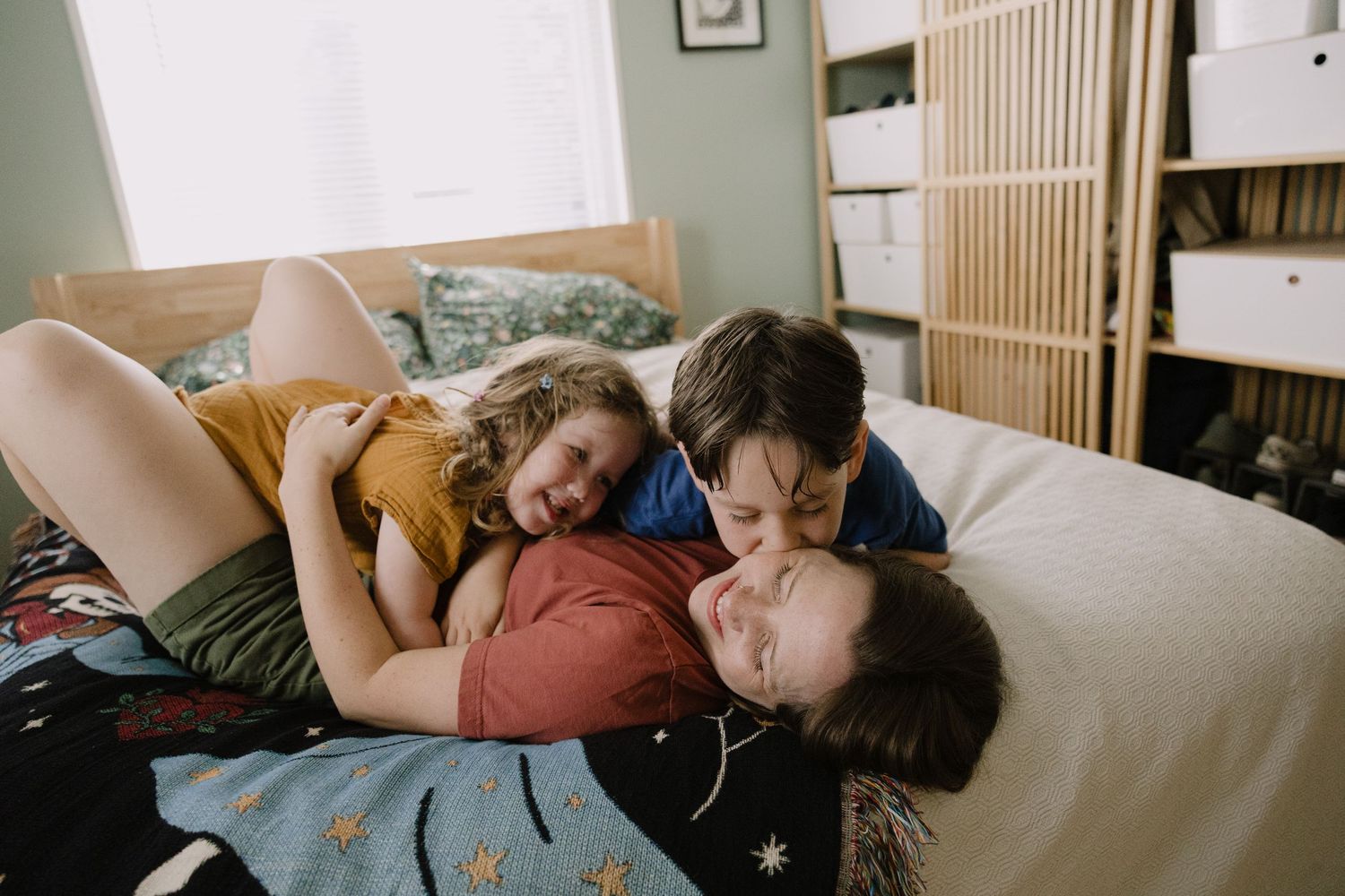 Sioux City, Iowa photographer. A mother lying on a bed laughing and cuddling with her two young children in a cozy bedroom.